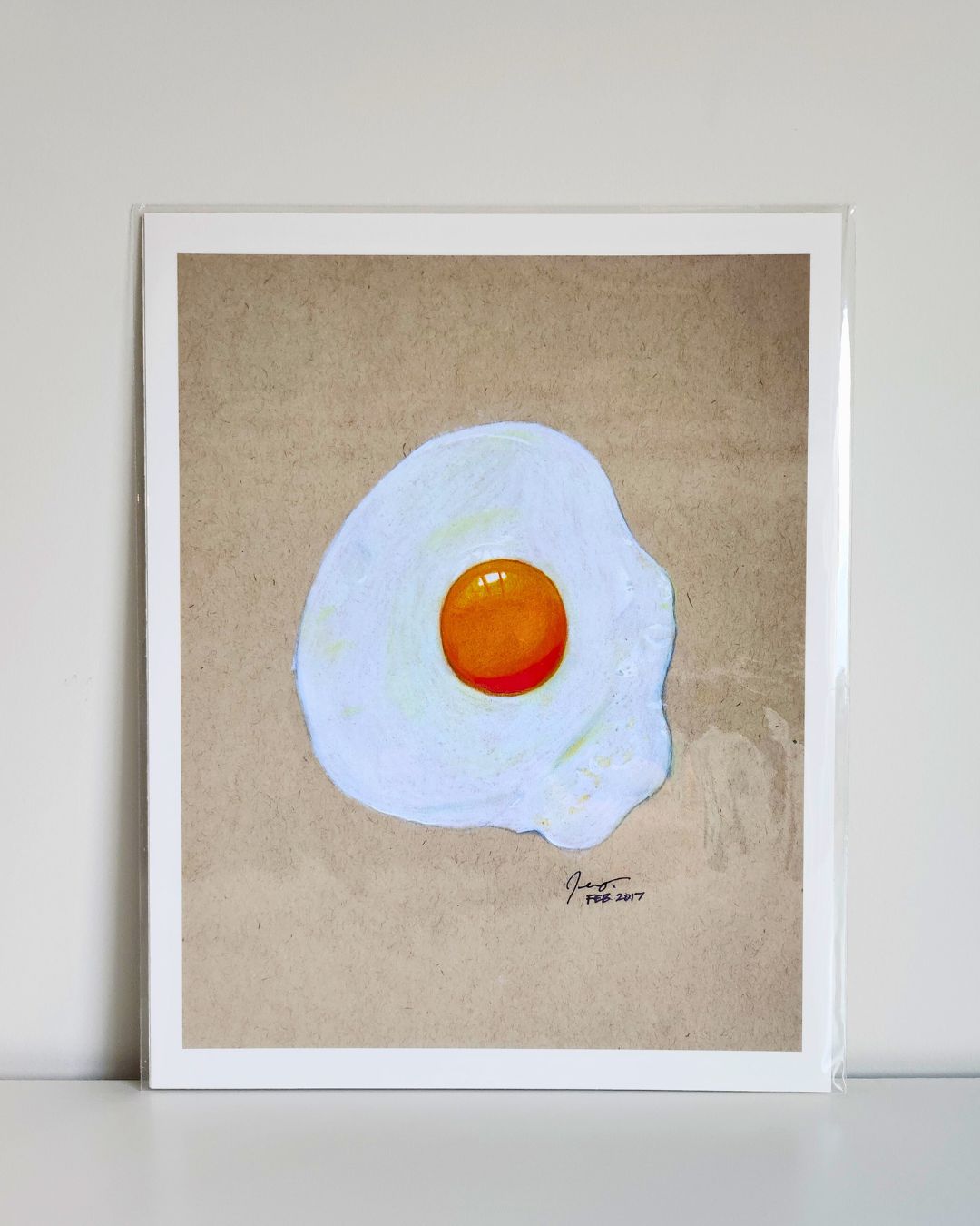 Fried egg realism art print by Full Art Alchemist, sleeved and displayed upright against white wall