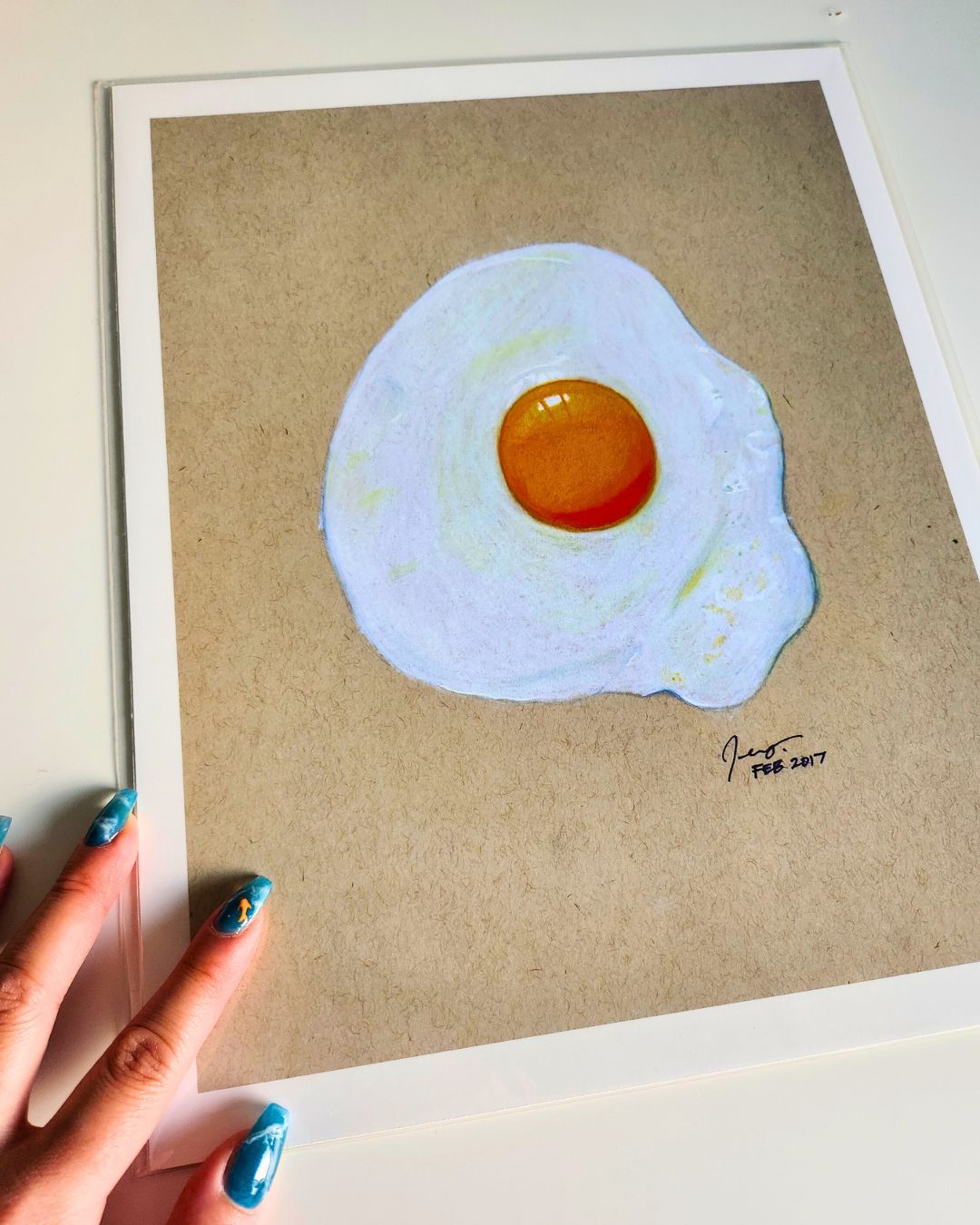 Fried egg print drawn in pencil crayon, photographed at an angle in archival sleeve with natural light