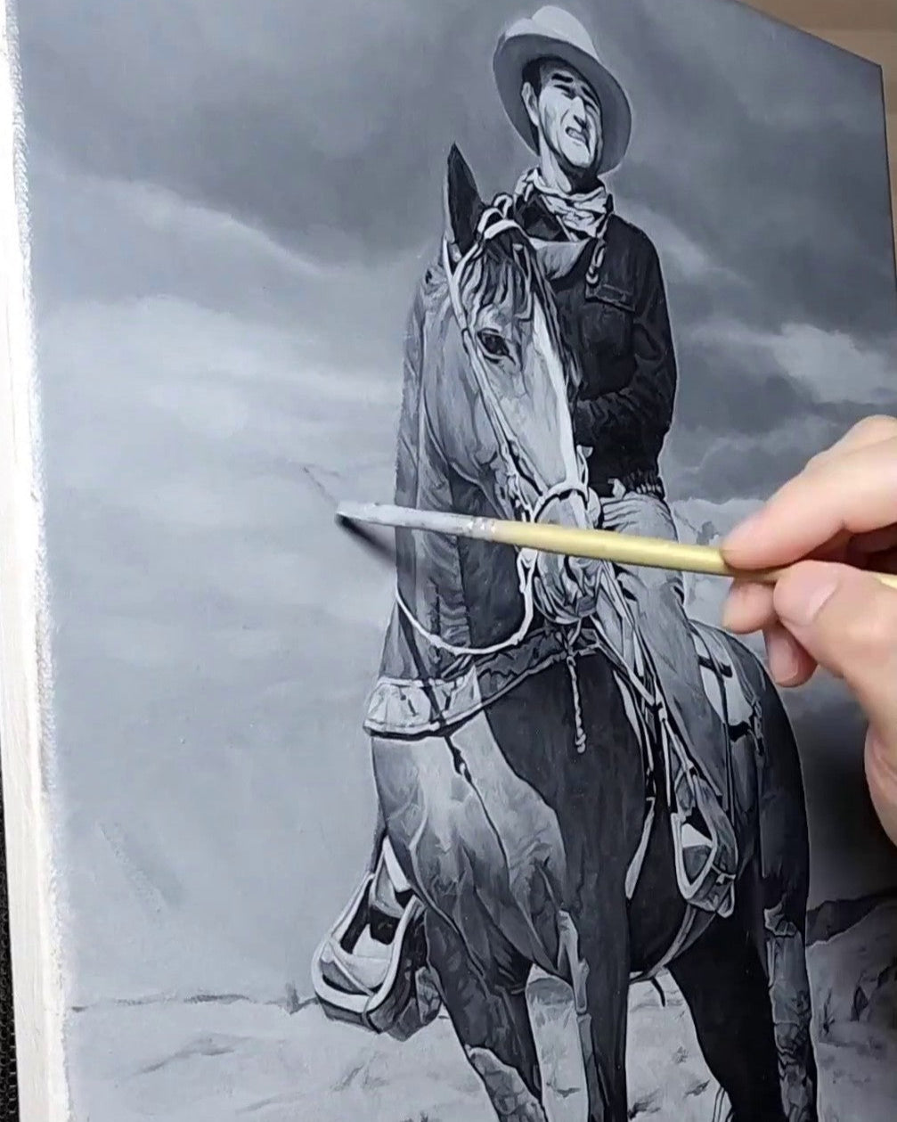 Black and white oil painting in progress of a cowboy on horseback. Artist's hand holds a paintbrush mid-stroke.
